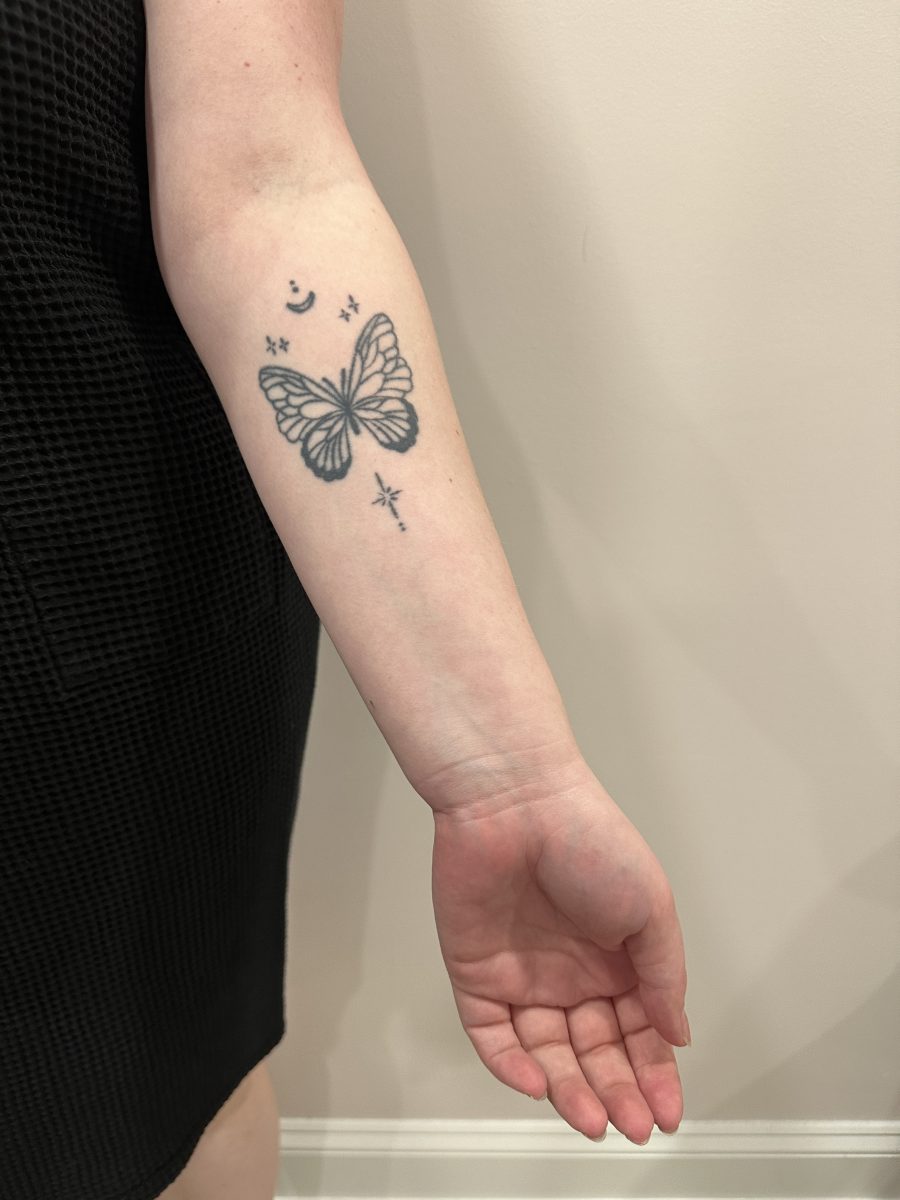 Forearm and hand showing natural light skin tone with a butterfly tattoo visible, before spray tan application by Liberty Greenwood at K Bella Hair Studio & Spa in Brighton, MI
