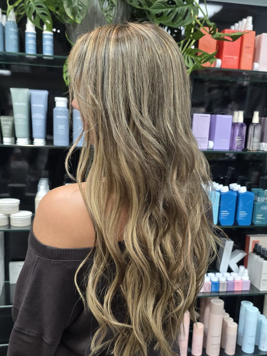 Long wavy dark blonde to brunette hair with fusion extensions, shown from behind, by Kristan Sayers at K Bella Hair Studio & Spa in Brighton, MI