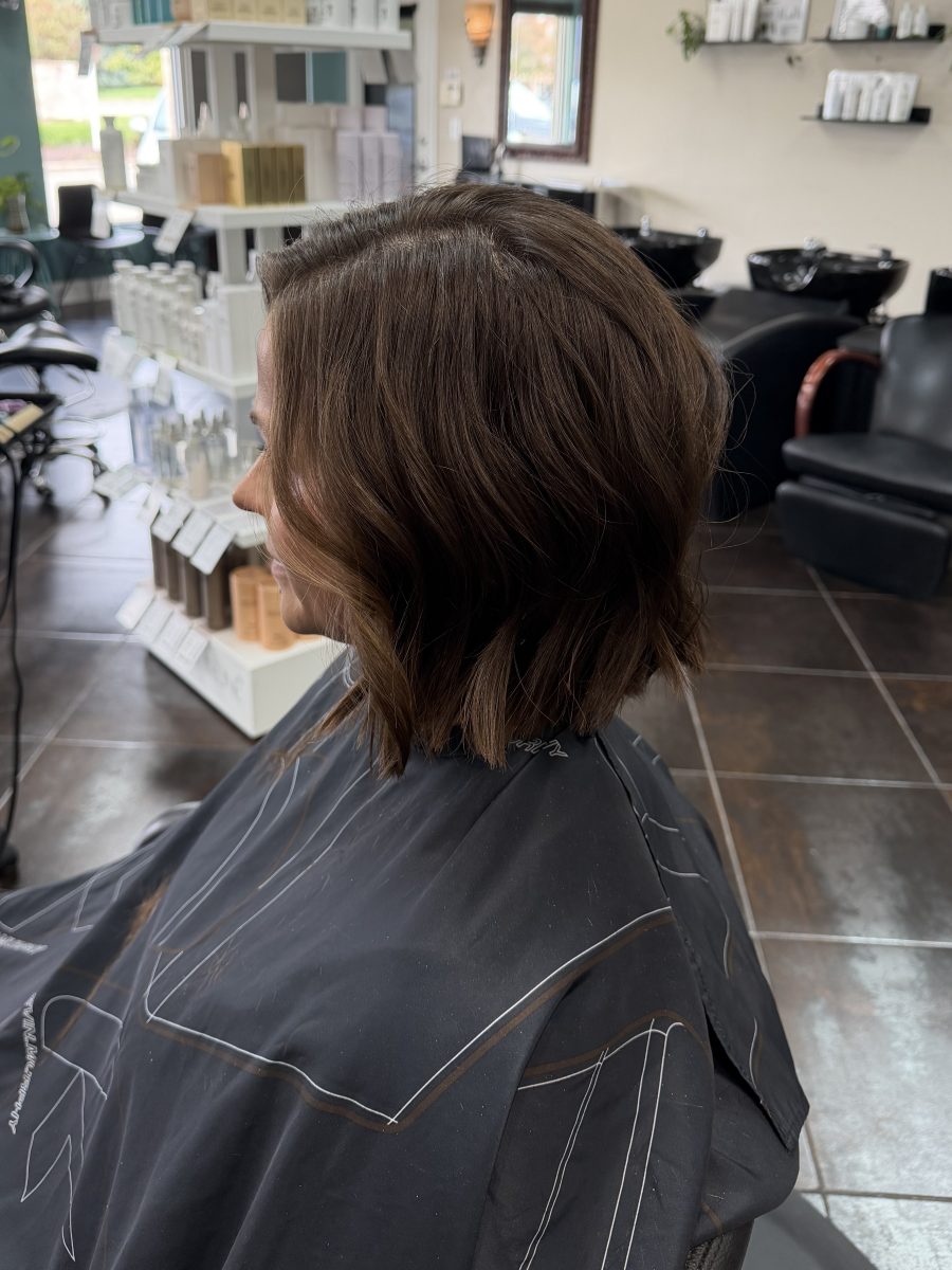 Back view of a woman with a shoulder-length textured haircut showing layered movement, wearing a black salon cape, by Katie Rowe at K Bella Hair Studio & Spa in Brighton, MI