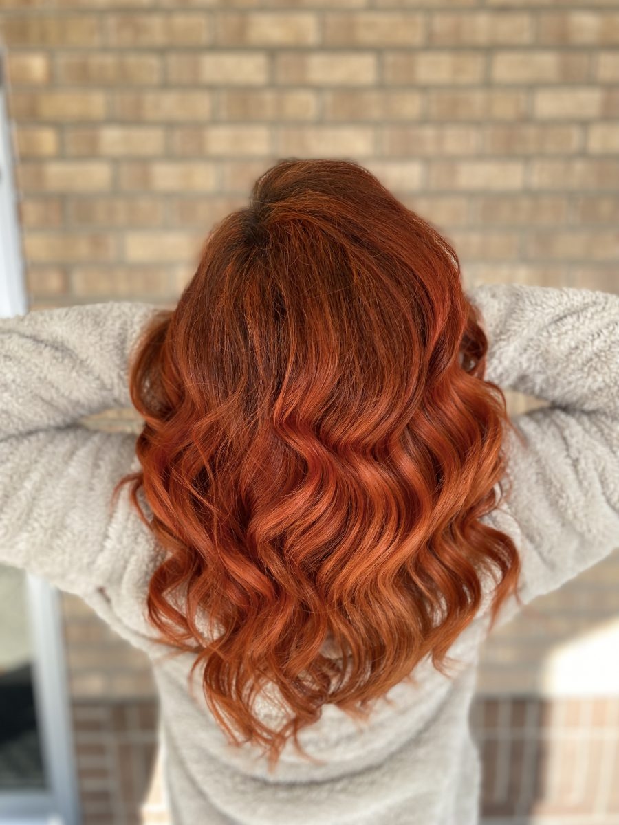 Back view of long wavy copper-toned hair with dimensional warmth, styled against a brick wall background by Allison Bowsher at K Bella Hair Studio & Spa in Brighton, MI